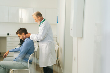 Side view of mature adult male manual therapist turning and bending head of male patient to check...