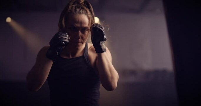 A Strong Fierce Kickboxing Woman Training On A Punching Bag In An MMA Gym. Shot In A Crossfit Boxing Gym With Low Key Lighting And A Scattering Of Haze. Captured On Red Digital Cinema Camera 