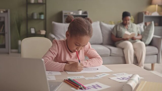 Waist up slowmo of 5 year old African-American gen A girl studying letters and coloring in coloring pages sitting at desk at home while her mother reading book in background
