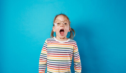 a little girl in big glasses and a striped T-shirt stands on a blue isolated background with her...