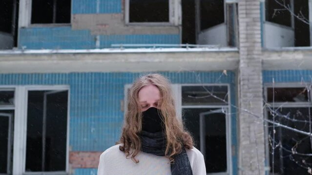 Young man wearing long blond hair and  black mask stands in front of  abandoned ruined building. Winter day, falling snow