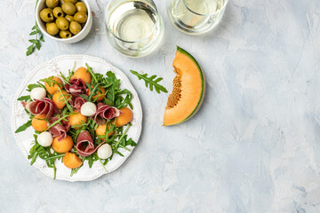 Fototapeta na wymiar mediterranean salad with prosciutto or jamon, mozzarella and green basil leaves and Cantaloupe melon on white table, traditional Spanish and Italian appetizer served with wholemeal grissini, top view