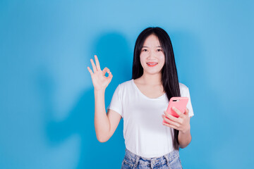 Smiling cheerful asian woman wears white t-shirt showing ok-sign holding mobile phone or smartphone on blue background