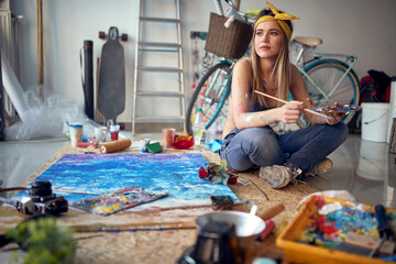Fototapeta premium A sexy and attractive female artist is thoughtfully sitting on the floor in her studio and waiting for the inspiration while working on the painting. Art, painting, studio