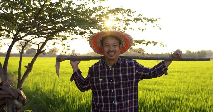 Slow motion Happy Asian Farmer holding a hoe and looking at camera green paddy field on background.