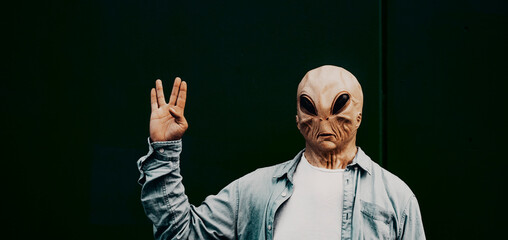 Alien mask people against a black background doing hallo gesture with hand. Portrait of extraterrestrial with human casual clothes. Banner header for immigration concept and invasion