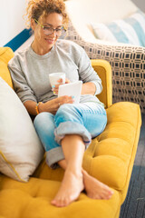 Confident caucasian adult woman sitting on the sofa reading a book using electronic device tablet...