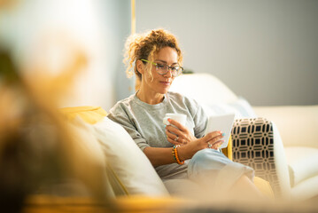 Relaxed young adult caucasian woman read a book on modern device reader comfortably sitting on the couch at home. Portrait of pretty female people wearing eyeglasses and using connection - Powered by Adobe