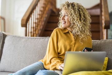 Cheerful adult lady with blonde long curly hair work at home on laptop computer comfortably sitting...