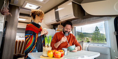 Young adult couple man and woman enjoy time inside a camper van cooking and speaking together with...