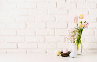 Fresh spring tulips in vase, gypsophila and pastel colored eggs on white brick wall background, copy space