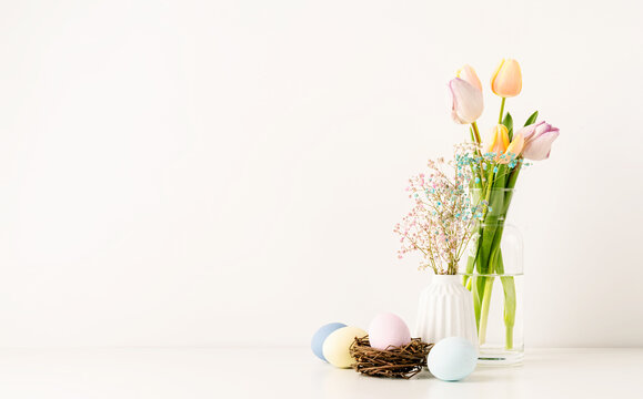 Fresh spring tulips in vase, gypsophila and pastel colored eggs on white wall background, copy space
