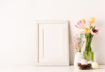 Mockup with a white frame spring tulips in vase, gypsophila and pastel colored eggs on light background