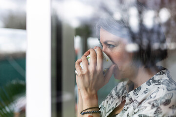 Close up of adult woman drinking coffee. She is at home looking outside through the window.