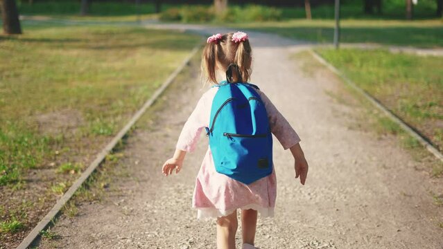 back to school. a little girl with a backpack runs through the green park to school along the path. education kid dream concept. a small child with a backpack back view runs the park to sun school