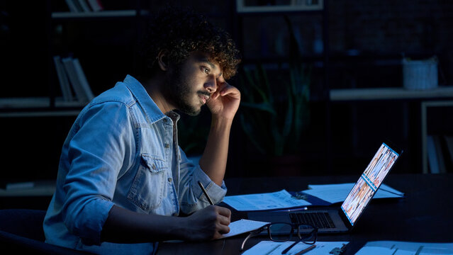 Focused Indian Latin Business Man Having Remote Video Call Conference Team Meeting Working Online By Distant Videocall Web Chat Late At Night At Home In Dark Office Using Laptop Computer.