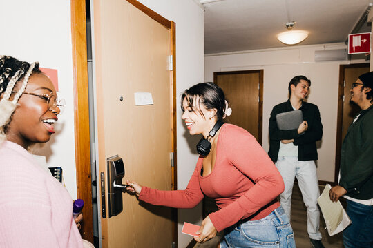 Happy young woman opening door while multiracial friends at corridor in college dorm