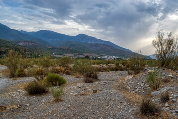 Old dried up river bed in a valley with hills behind and scrub grass and weeds