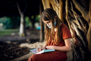 A cute european baby girl in a dress and a mask sits near a big tree in the park. A child among the big roots of a tree writes poetry, a girl makes notes