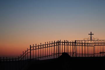 A fence with a religious cross attached to it next to a church and the aegean sea, while the sun is setting in a dramatic way