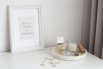 Frame with text BEAUTY IN SIMPLICITY, marble tray with perfume, jewelry and candles stand on white chest of drawers. Spring bedroom home decor. White stylish interior.