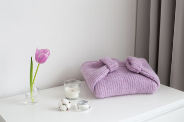 Fototapeta na wymiar Pink tulip in glass vase, lilac knitted sweater and candles stand on white chest of drawers. Spring bedroom home decor. White stylish interior.