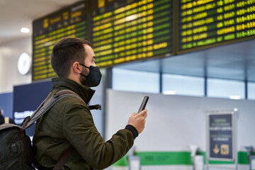 Young man with mask and backpack at the airport looking at flight schedule through mobile.