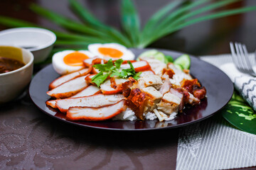 Steamed rice and red pork, crispy pork in a black plate Served with dipping sauce and dark soy sauce Place it on a black wooden table.