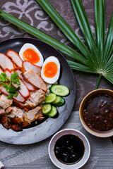 Steamed rice and red pork, crispy pork in a black plate Served with dipping sauce and dark soy sauce Place it on a black wooden table.
