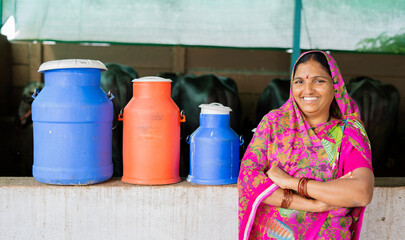 Portrait shot of happy Indian milk dairy woman farmer with milk containers and cattles behind...
