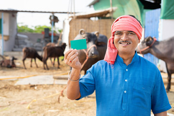 Smiling indian milk dairy farmer showing green card by looking at camera - concept of advertisement, promotions and successful - obrazy, fototapety, plakaty