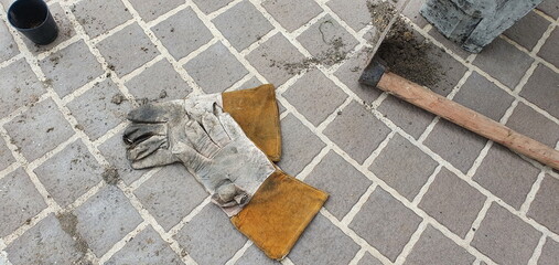 White faux leather gloves with sloppy brown edges. And a wooden-handled iron shovel was placed on a rectangular tiled floor on a garden path with small black plastic plant pots nearby.
