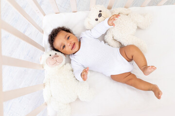 Smiling African-American little baby in white sleeping bed with bear toys