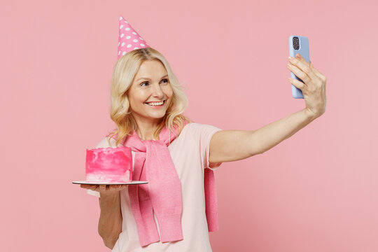 Elderly Happy Smiling Woman 50s Wear T-shirt Birthday Hat Hold Cake Doing Selfie Shot On Mobile Cell Phone Isolated On Plain Pastel Pink Background Studio Portrait. Celebration Party Holiday Concept.