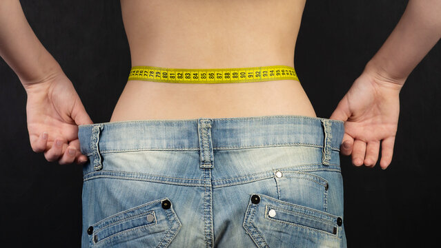 Fit, beautiful, young girl in oversized pants close-up. A thin girl with weight loss shows a flat stomach, pulling big jeans with her hands.The concept of diet and weight loss. Black background.