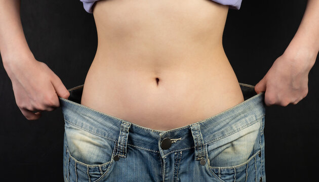 Fit, beautiful, young girl in oversized pants close-up. A thin girl with weight loss shows a flat stomach, pulling big jeans with her hands.The concept of diet and weight loss. Black background.