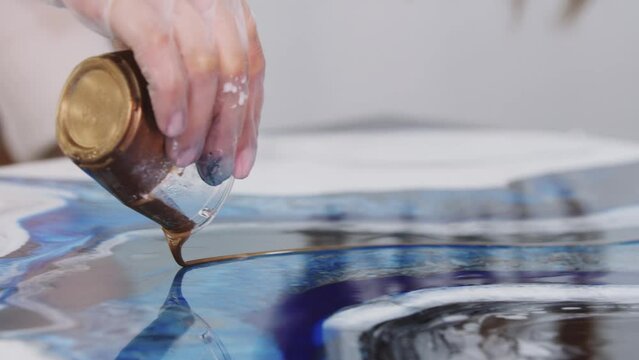 Young woman pouring down the golden glittery epoxy on the painting from the cup