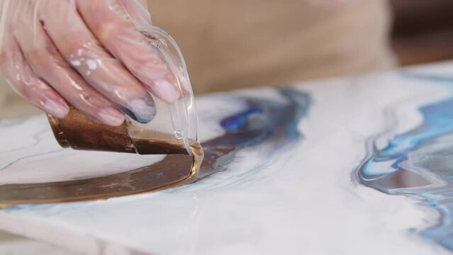 Young woman pouring down the golden glittery epoxy on the painting from the plastic cup
