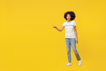 Full body young woman of African American ethnicity in white volunteer t-shirt walk go point finger aside workspace isolated on plain yellow background. Voluntary free assistance help grace concept.