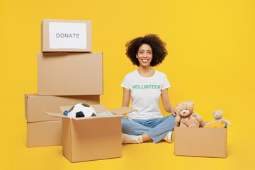 Full body smiling young woman of African American ethnicity wear white volunteer t-shirt sit near boxes with presents isolated on plain yellow background. Voluntary free work assistance help concept
