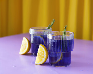 Tea with ice from clitoria flowers in transparent glasses on a lilac table. Slices of lemon next to the glasses. Sprigs of rosemary in the background. Yellow background