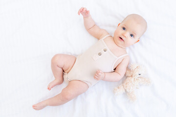 a newborn baby baby girl in a cotton suit on a white bed at home is lying and smiling with a teddy bear