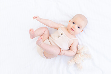 a newborn baby baby girl in a cotton suit on a white bed at home is lying and smiling with a teddy bear