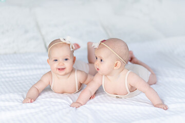 two newborn baby twin girls in a cotton suit on a white bed at home lie on their stomach and smile...
