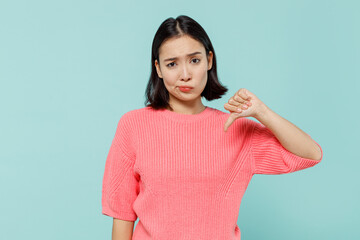 Fototapeta premium Young sad displeased unhappy woman of Asian ethnicity 20s wear pink sweater showing thumb down dislike gesture isolated on pastel plain light blue background studio portrait. People lifestyle concept.
