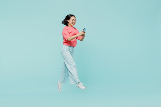 Full Body Young Smiling Happy Woman Of Asian Ethnicity 20s Wearing Pink Sweater Jump High Hold In Hand Mobile Cell Phone Isolated On Pastel Plain Light Blue Color Background. People Lifestyle Concept.