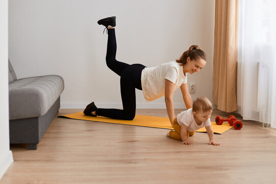 Image of positive adorable woman wearing white t shirt and black leggins doing sport exercises at home, doing one legged plank pose bow leg with her baby, training muscle flexibility.