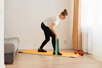 Portrait of slim attractive woman wearing white t shirt and black leggins doing sport exercises at home, standing with bending upper body and holding resistance band for workout.