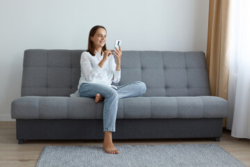 Indoor shot of happy Caucasian beautiful woman holding mobile phone while sitting on a sofa, chatting with friends, wearing white shirt and jeans, expressing positive emotions.