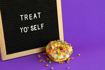 A fruity rice cereal topped donut with a letterboard sign that says "Treat Yo'self" on a bright purple background